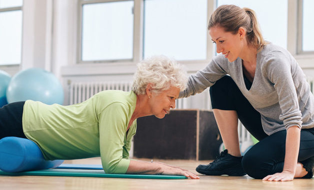 photodune-10122819-physical-therapist-helping-elderly-woman-in-her-workout-s-1 Senior woman exercising with a foam roller being assisted by personal instructor at gym. Physical therapist helping elderly woman in her workout at health club.